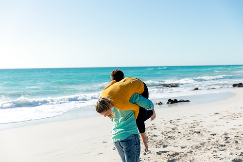 Rear view of a Caucasian couple standing on the beach with blue sky and sea in the background, piggybacking and having fun