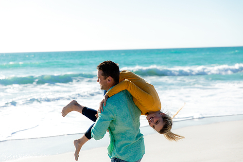 Side view of a Caucasian man carrying his Caucasian girlfriend over his shoulder, walking on the beach with blue sky and sea in the background, smiling