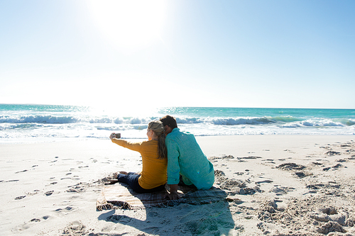Rear view of a Caucasian couple reclining on the beach with blue sky and sea in the background, embracing and taking selfie