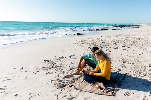 Side view of a Caucasian couple reclining on the beach with blue sky and sea in the background, embracing and looking at their smartphone