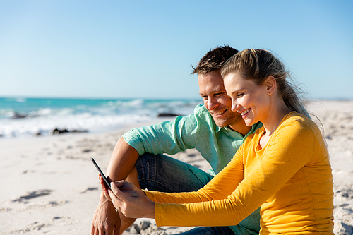 Side view of a Caucasian couple reclining on the beach with blue sky and sea in the background, embracing and looking at their smartphone