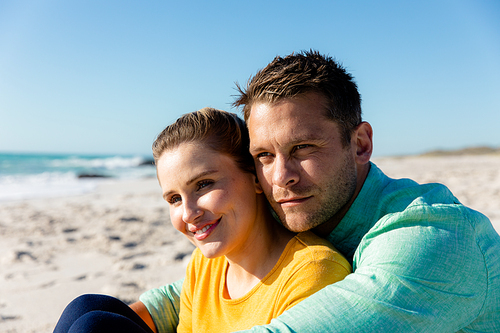 Front view of a Caucasian couple reclining on the beach with blue sky and sea in the background, embracing and looking away