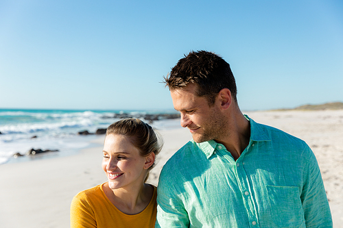 Front view of a Caucasian couple walking on the beach with blue sky and sea in the background, smiling and looking at each other