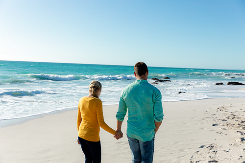 Rear view of a Caucasian couple walking on the beach with blue sky and sea in the background, holding hands