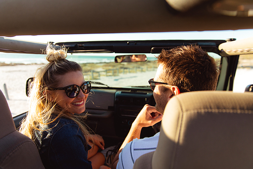 Rear view of a Caucasian couple sitting inside an open top car with blue sky and sea in the background, the man driving and the woman smiling to camera