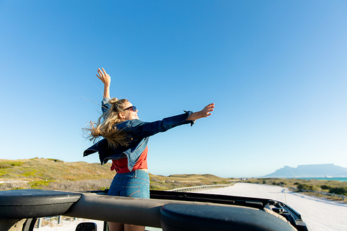 Rear high angle view of a Caucasian woman inside an open top car, standing, smiling and raising her hands. Weekend beach vacation, lifestyle and leisure.