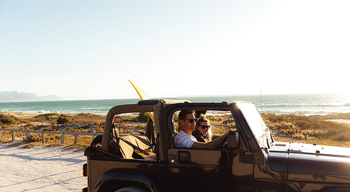 Side view of a Caucasian couple inside an open top car, with blue sky and sea in the background, driving and smiling