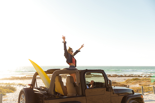 Side view of a Caucasian woman inside an open top car, smiling, standing and raising her hands. Weekend beach vacation, lifestyle and leisure.