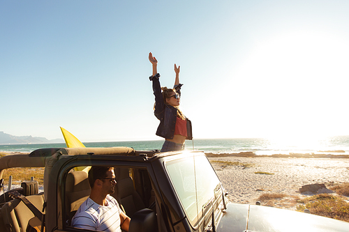 Side view of a Caucasian couple inside an open top car, the man driving, the woman smiling, standing and raising her hands