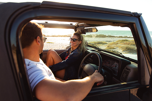 Side view of a Caucasian couple inside an open top car, talking and smiling at each other. Weekend beach vacation, lifestyle and leisure.