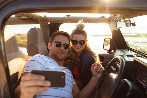 Front view of a Caucasian couple inside an open top car, smiling and taking selfie. Weekend beach vacation, lifestyle and leisure.