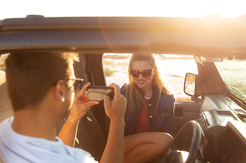 Rear view of a Caucasian man with his girlfriend smiling inside an open top car, taking a photo of her