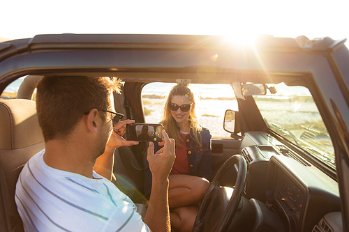 Side high angle view of a Caucasian man with his girlfriend smiling inside an open top car, taking a photo of her