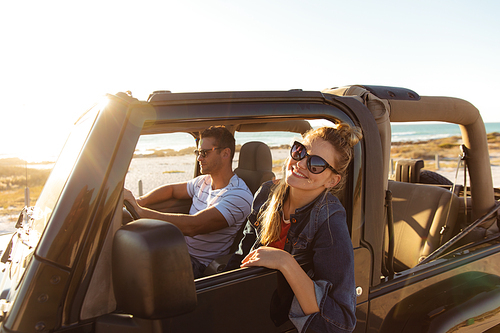 Side view of a Caucasian couple inside an open top car, the man driving, the woman smiling and looking through the window