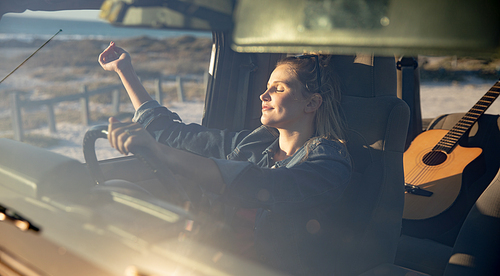 Front view of a Caucasian woman inside an open top car, holding a steering wheel, raising her hands and relaxing in the sun with eyes closed