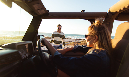 Side view of a Caucasian couple, the woman inside an open top car holding a steering wheel, the man looking at his partner