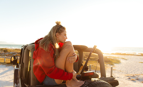 Side view of a Caucasian woman sitting on the tyre of an open top car, looking away. Weekend beach vacation, lifestyle and leisure.
