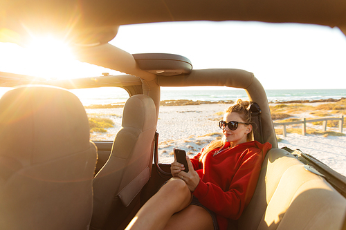 Front view of a Caucasian woman sitting inside an open top car, using her mobile phone and smiling. Weekend beach vacation, lifestyle and leisure.