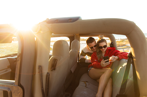 Front view of a Caucasian couple inside an open top car, using their smartphone and smiling. Weekend beach vacation, lifestyle and leisure.
