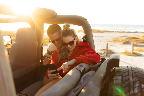 Front view of a Caucasian couple inside an open top car, using their smartphone and smiling. Weekend beach vacation, lifestyle and leisure.