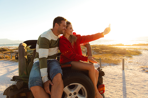 Front view of a Caucasian couple sitting on a tyre of an open top car, smiling and taking selfie. Weekend beach vacation, lifestyle and leisure.