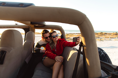 Front view of a Caucasian couple inside an open top car, smiling and taking selfie. Weekend beach vacation, lifestyle and leisure.
