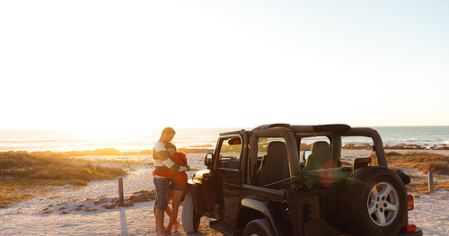 Front view distant of a Caucasian couple next to an open top car, embracing and smiling. Weekend beach vacation, lifestyle and leisure.