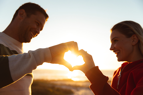 Front view of a Caucasian couple, smiling at each other and making a shape of heart with their hands . Weekend beach vacation, lifestyle and leisure.