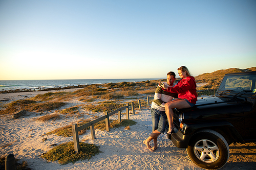 Front view of a Caucasian couple outside their open top car, embracing and looking at the sea. Weekend beach vacation, lifestyle and leisure.