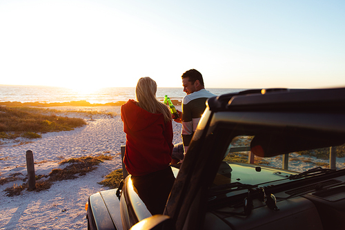 Rear view of a Caucasian couple outside their open top car, with sunset on the beach in the background, making a toast