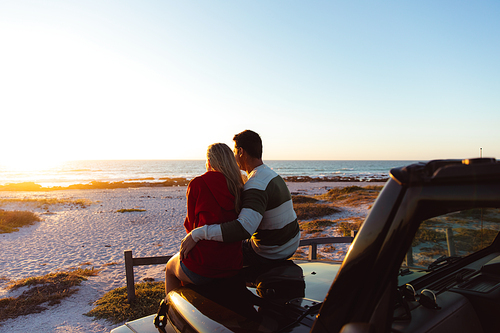 Rear view of a Caucasian couple outside their open top car, with sunset on the beach in the background, embracing and looking away