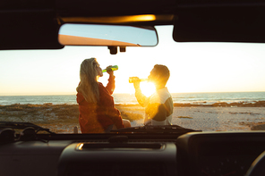 Rear view of a Caucasian couple outside their open top car, with sunset on the beach in the background, drinking beer