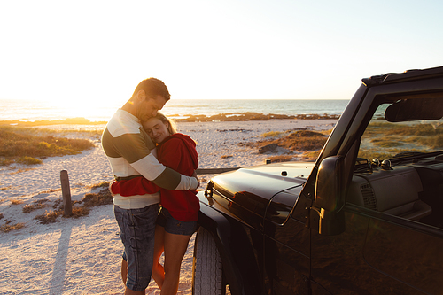 Front view of a Caucasian couple outside their open top car, with sunset on the beach in the background, embracing and smiling