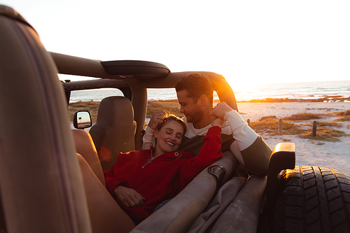 Front view of a Caucasian couple inside an open top car, with sunset on the beach in the background, embracing, smiling, holding hands and having fun