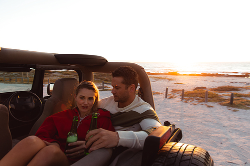 Front view of a Caucasian couple inside an open top car, with sunset on the beach in the background, embracing, talking and holding bottles of beer