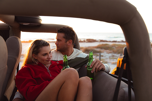 Front view of a Caucasian couple inside an open top car, with sunset on the beach in the background, embracing and holding bottles of beer