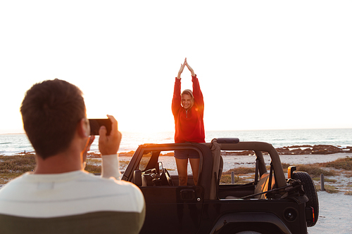 Front view of a Caucasian couple outside an open top car, with sunset on the beach in the background, the man taking a photo of his partner raising hands and smiling