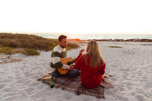 Rear view of a Caucasian couple reclining on the beach, with sunset in the background, the man holding the guitar, the woman holding food