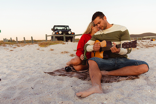 Front view of a Caucasian couple reclining on the beach, with an open top car in the background, the man holding the guitar, the woman embracing his partner
