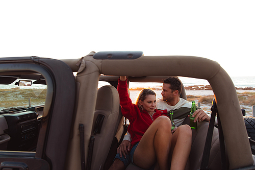 Front view of a Caucasian couple inside an open top car, with sunset on the beach in the background, embracing and holding bottles of beer