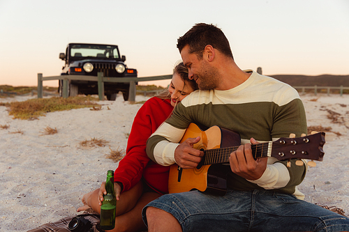 Front view of a Caucasian couple reclining on the beach, with an open top car in the background, the man holding the guitar, the woman hugging her partner and holding a bottle of beer