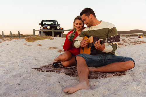 Front view of a Caucasian couple reclining on the beach, with an open top car in the background, the man holding the guitar, the woman holding a bottle of beer