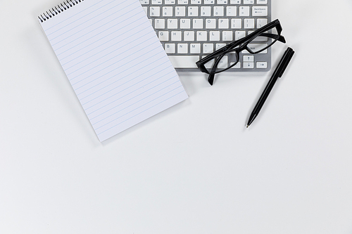 Close up top view of an empty page in a notebook, a pen, a keyboard and glasses arranged on a plain white background