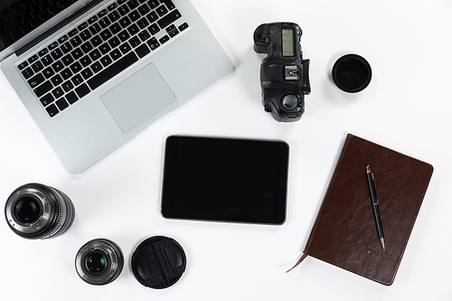 Close up top view of a notebook, a pen, a laptop, a tablet, a camera and lenses in various sizes arranged on a plain white background