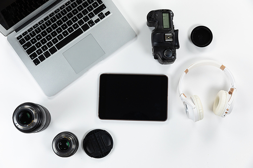 Close up top view of headphones, a laptop, a tablet, a camera and lenses in various sizes arranged on a plain white background