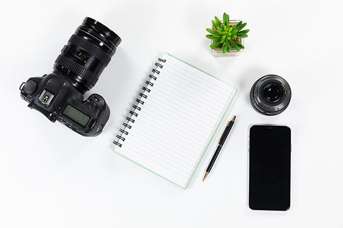 Close up top view of a notebook, a pen, a smartphone, a camera and a plant arranged on a plain white background