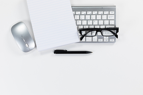 Close up top view of an empty page in a notebook, a pen, a keyboard, a computer mouse and glasses arranged on a plain white background