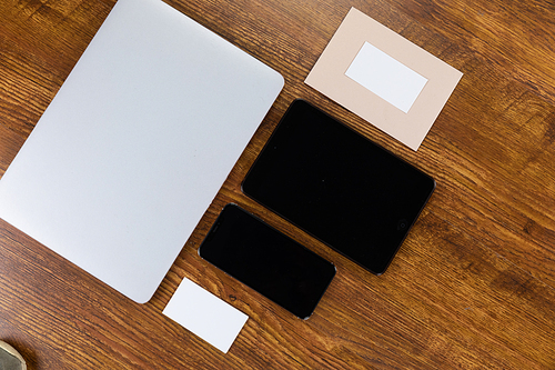 Close up top view of a laptop, a tablet, a smartphone and sheets of paper in various sizes arranged on a textured wooden surface