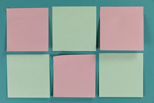 Close up top view of six green and pink sheets of paper sticky memo notes in one size, arranged on a plain blue background