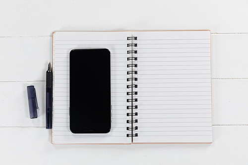 Close up top view of a black smartphone, a notebook and a black pen arranged on a plain white background